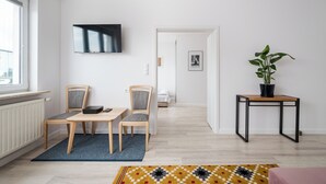 A modern living room with a TV mounted on the wall, a dining area with a table and chairs, a potted plant on a side table, and a view into another room through an open door.