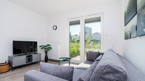 A modern living room with a grey sofa, a flat-screen TV, a potted plant, and a view of buildings through large windows.