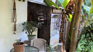 Ein überdachter Außenbereich mit Tisch und Stühlen, einer hängenden Pflanze und einer Holzhütte im Hintergrund.
