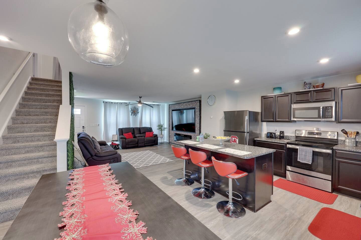 A modern apartment with a kitchen, dining area, and living room. It includes a staircase, a sofa, a TV, and a kitchen island with bar stools.