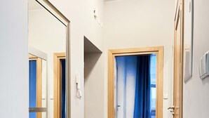 A narrow hallway with wooden flooring, a large mirror, and a door leading to a bedroom with a bed and a window with blue curtains. - Welcoming Apartment with two bedrooms, Świętego Jana (Unknown)