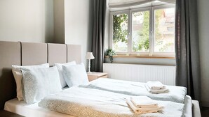 A neatly made bed with white linens and a patterned blanket, a bedside table with a lamp, and a window with curtains.