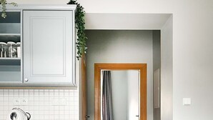 A modern kitchen with white cabinets, a coffee machine, and a wooden countertop. A hallway leads to a bedroom with a bed and a window.