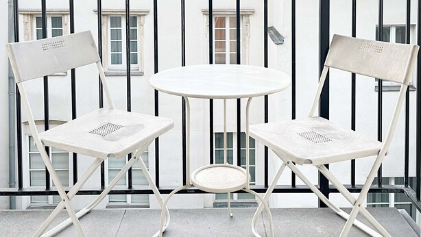 A balcony with two white folding chairs and a small round table, overlooking a building with a grey roof and white walls.