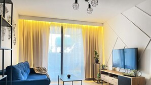 Ein modernes Wohnzimmer mit einem blauen Sofa, einem Glastisch, einer Holz-TV-Konsole und einem großen Fenster mit gelben Vorhängen.