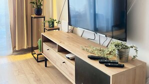 Ein modernes Wohnzimmer mit einem großen Flachbildfernseher, der an einer Holzkonsole befestigt ist, einem kleinen Beistelltisch mit einem Topfblumen und einem Fenster mit transparenten Vorhängen.