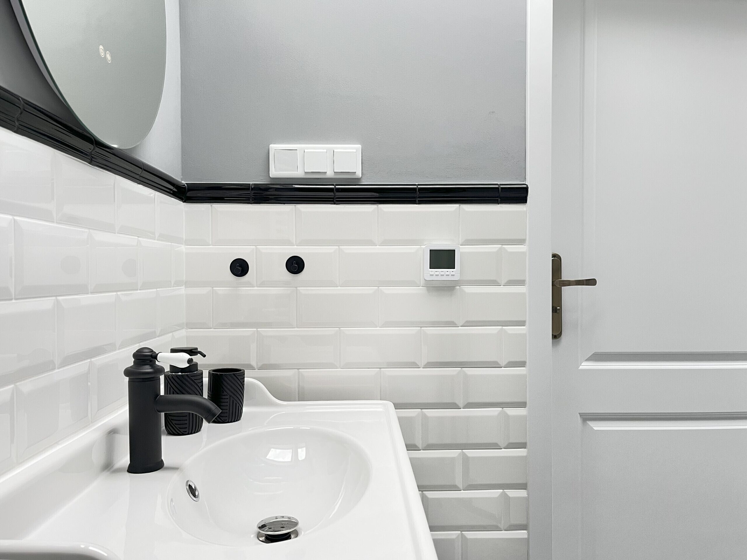 A modern bathroom with a white sink, black faucet, and a digital thermostat on the wall.