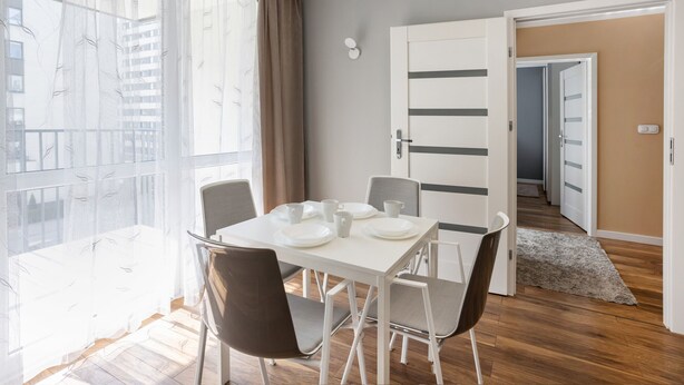A dining area with a table set for four, a window with sheer curtains, and a door leading to another room.