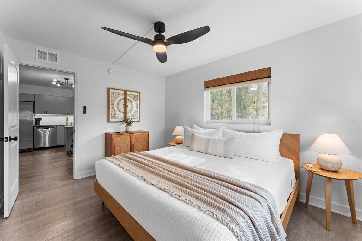 A bedroom with a bed, bedside tables, a ceiling fan, a window, and a view of a kitchen.