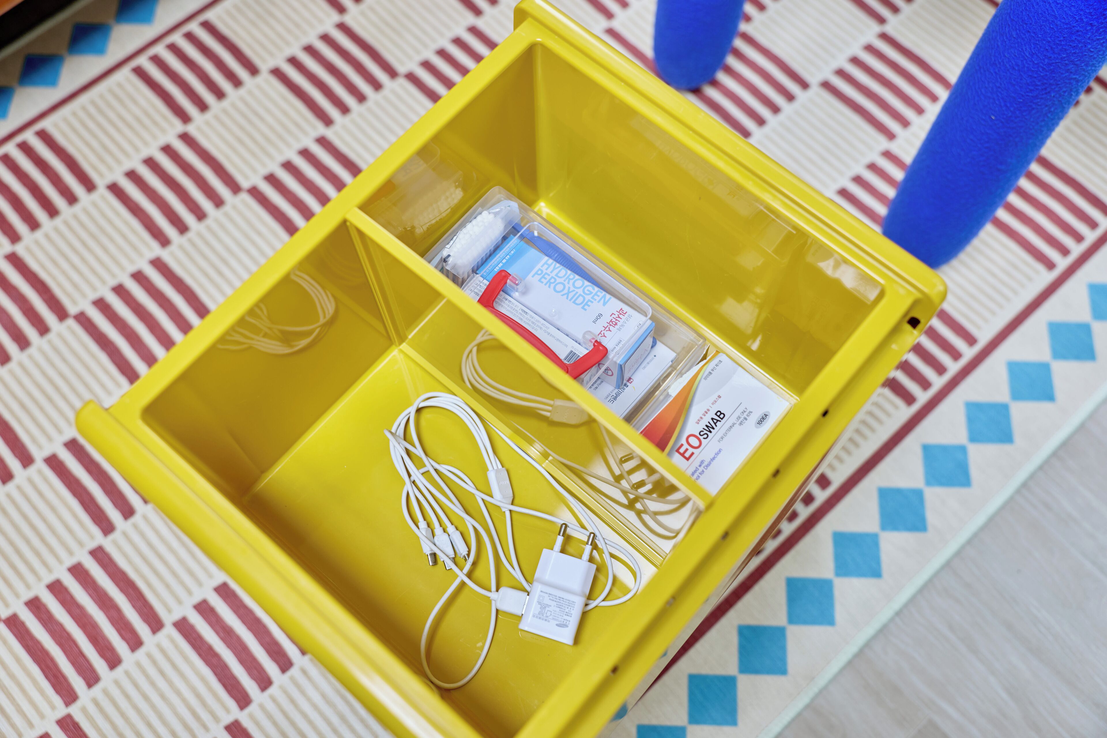 Une boîte de rangement jaune contenant des câbles et une boîte de peroxyde d’hydrogène, le tout posé sur un sol à motifs.