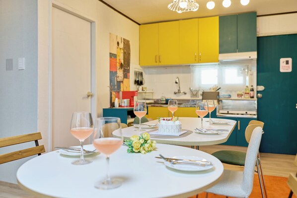 A dining area with round tables, chairs, and pink drinks. There is a kitchen in the background with yellow and teal cabinets.