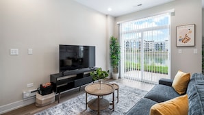 Una sala de estar moderna con un televisor de pantalla plana, un sofá, una mesa de centro y un amplio ventanal con vistas a edificios y zonas verdes.