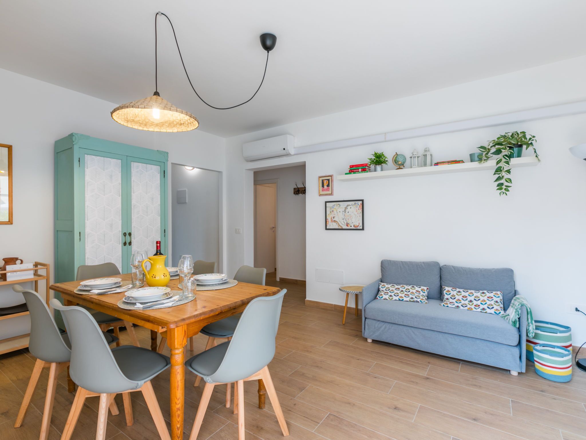 A modern living room with a wooden dining table, grey chairs, a blue sofa, and a hanging light fixture.