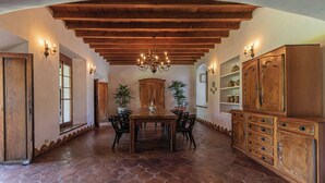 Un vaste espace dédié à la salle à manger, avec une grande table en bois, des chaises et un lustre. Des plantes en pot et des meubles en bois sont disposés le long des murs.