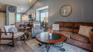 A spacious living room with a brown leather sofa, a round coffee table, and a grey armchair. A kitchen with a window and a clock is visible in the background. - 2BR Lake View Indian Point Condo Next to Silver Dollar City | (GF17-5) (Branson)
