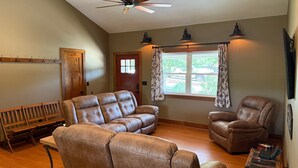 A spacious living room with a ceiling fan, leather sofas, a wooden bench, and a large window with curtains.