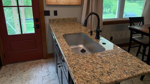 A kitchen with granite countertops, a stainless steel sink, a wooden door, and a window with a view of greenery.