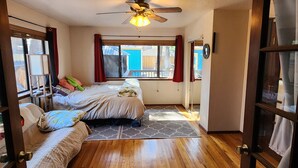 Un dormitorio con suelo de madera, una cama con muchas almohadas, un ventilador de techo y una ventana con cortinas rojas.