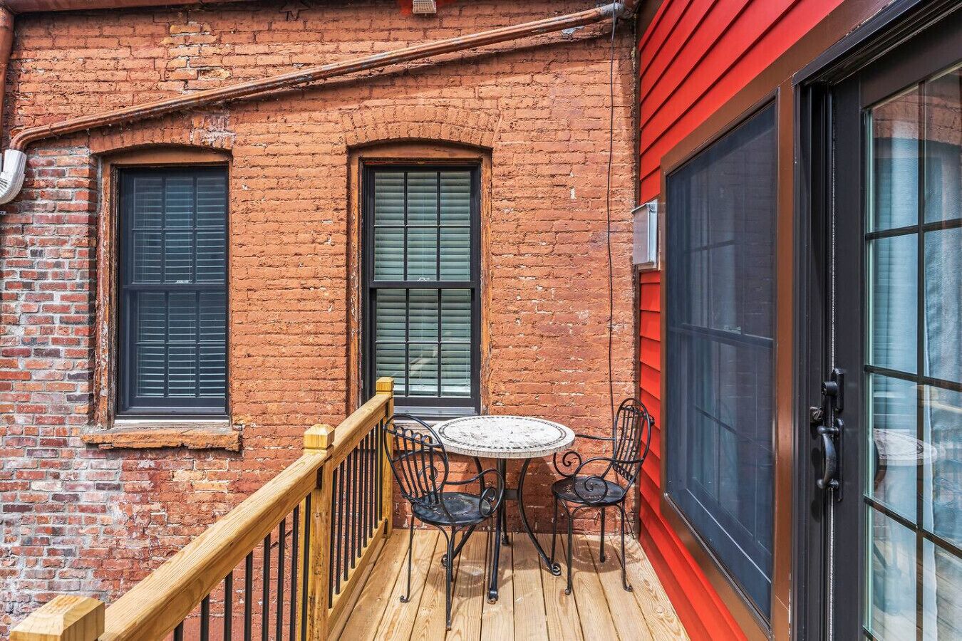 A brick building with a wooden deck, two windows, a table with chairs, and a red door.