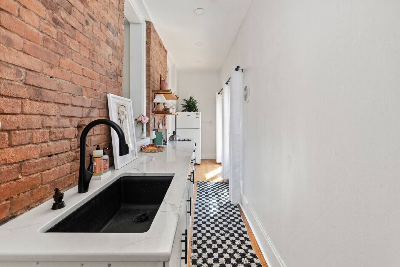 A narrow hallway with a kitchen area featuring a black and white checkered floor, a white sink with a black faucet, and a brick wall.