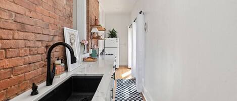 A narrow hallway with a kitchen area featuring a black and white checkered floor, a white sink with a black faucet, and a brick wall.