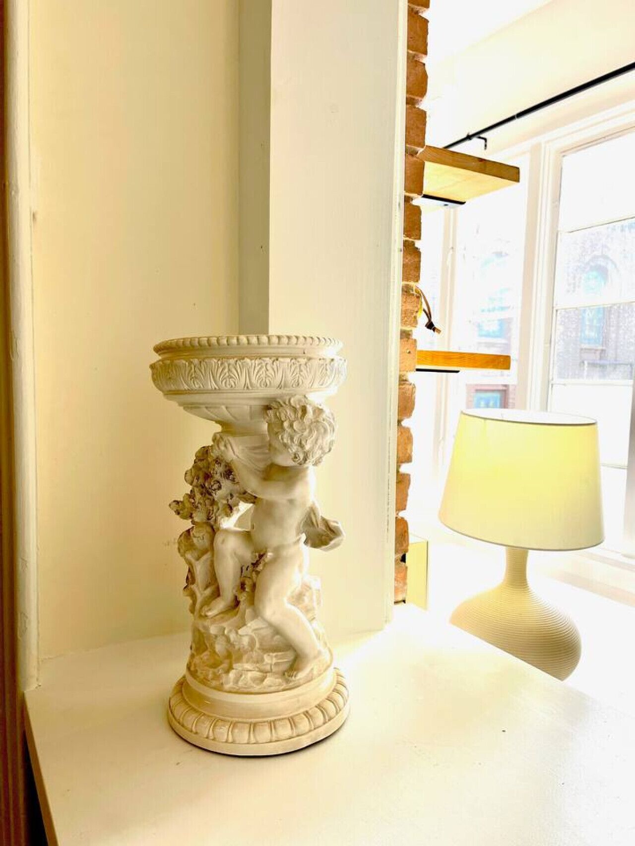 A white pedestal with a decorative base featuring a figure, a white table lamp, and a window with a view of buildings.