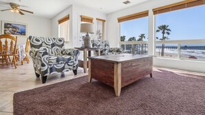 A spacious living room with a large wooden coffee table, patterned armchairs, and a view of the beach through large windows.