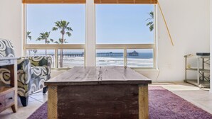 A room with a wooden table, a patterned armchair, and large windows overlooking a beach with palm trees and a pier.