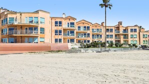 A beachfront hotel with multiple balconies, a palm tree, and a sandy beach in the foreground.