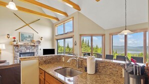 A kitchen with granite countertops, a stone fireplace, and large windows offering a view of trees and mountains.
