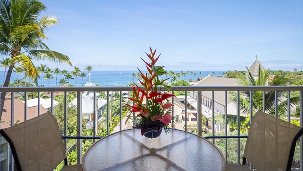 Un balcon avec une table et des chaises, offrant une vue sur la côte, avec des palmiers et une église.