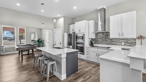 A modern kitchen with white cabinetry, a central island, and a dining area with a pool table.