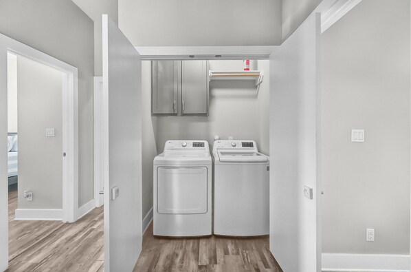 A laundry room with two front-loading washers and dryers, a small shelf above them, and a wooden floor. - Waterfront Escape | Rooftop Terrace + Private Pool + Boat Slips (Corpus Christi)