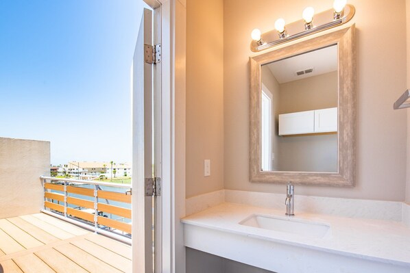 A bathroom with a white sink, a mirror with lights, and a view of a balcony with a railing.
