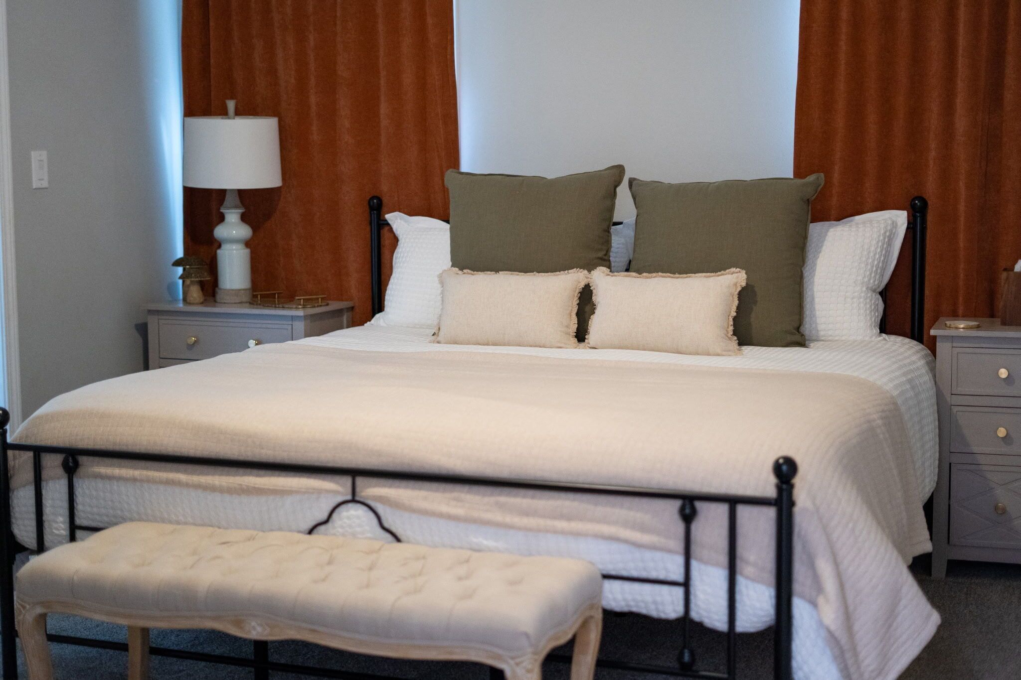 A neatly made bed with a white comforter and two pillows, a bedside table with a lamp, and a bench at the foot of the bed.