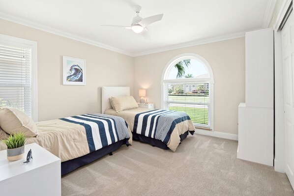 A bedroom with two beds, a ceiling fan, a window with a view of greenery, and a white wardrobe.