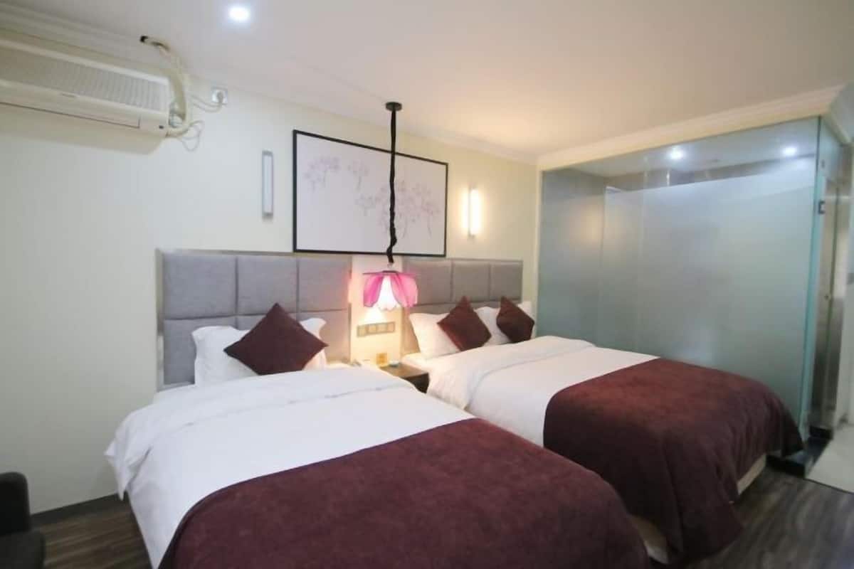 Une chambre d’hôtel avec deux lits, une table de chevet avec une lampe, un climatiseur fixé au mur et une porte en verre donnant sur la salle de bain.