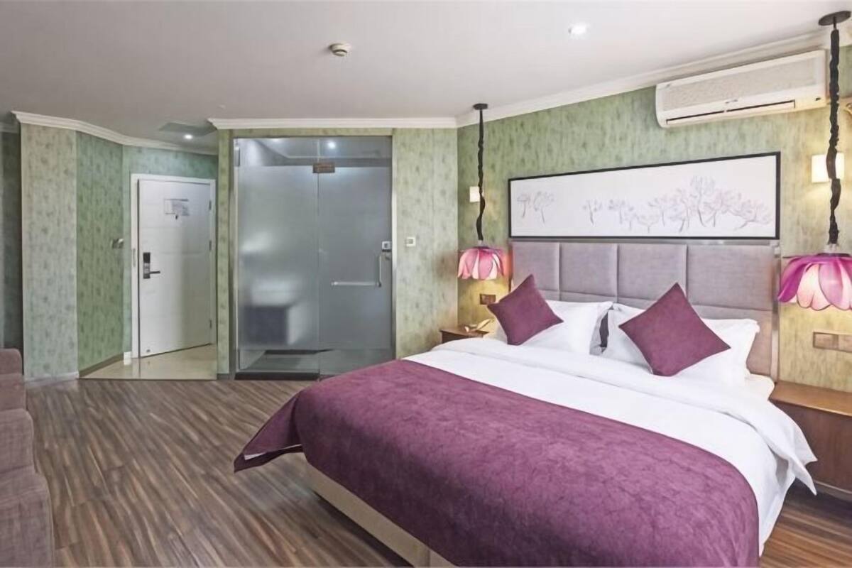 Une chambre d’hôtel avec un lit, une table de chevet, un climatiseur fixé au mur, une porte et un couloir.