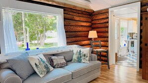 A cozy log cabin interior with a grey sofa, a wooden chair, and a view of the outdoors through a large window.