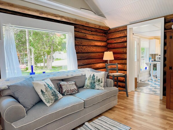 A cozy log cabin interior with a grey sofa, a wooden chair, and a view of the outdoors through a large window.