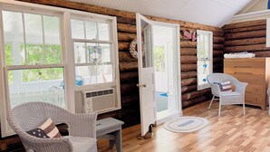 A log cabin interior with a wicker chair, a wooden dresser, and a large window with a stained glass design.