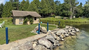 A lakeside cabin with a small deck, surrounded by a rocky shoreline and a grassy area.