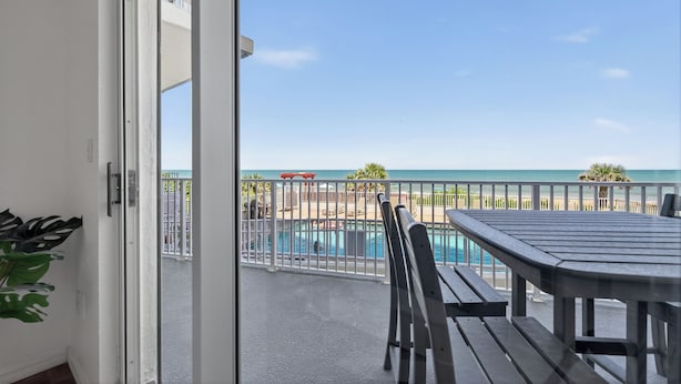 Un balcon avec vue sur une piscine et une plage, une table et des chaises.