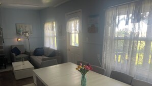 A room with a white dining table, a sofa, and a chair. There are windows with curtains, a painting on the wall, and a vase with flowers on the table.