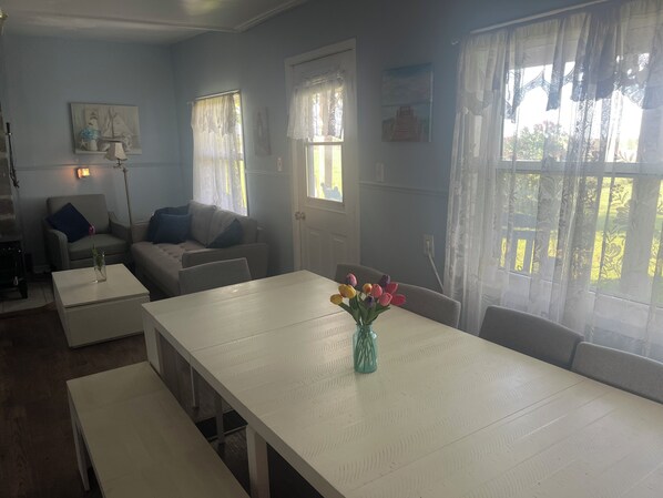 A room with a white dining table, a sofa, and a chair. There are windows with curtains, a painting on the wall, and a vase with flowers on the table. - Sun and Stars farmhouse, North Cape Coastal drive (Richmond)