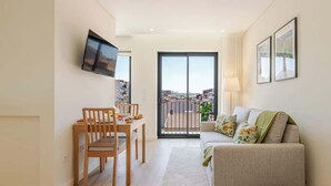 A modern living room with a sofa, a dining table with chairs, a TV mounted on the wall, and a balcony with a view of the city.