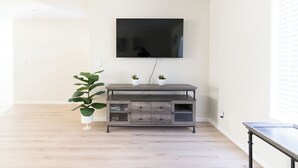 A modern living room with a flat-screen TV mounted on the wall, a dark wooden entertainment unit with two potted plants, and a large potted plant to the left.