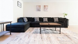 A modern living room with a sectional sofa, a coffee table, and a rug. There are two framed artworks on the wall and a small plant on the right.