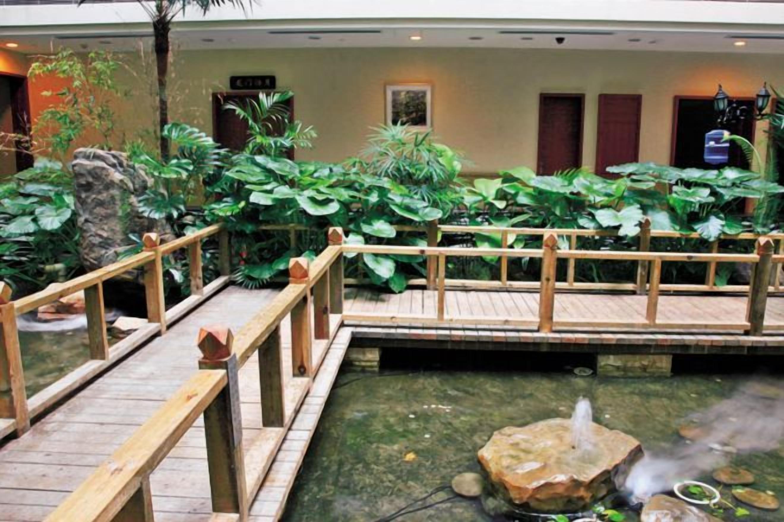 A wooden bridge over a pond with a fountain, surrounded by lush greenery and a wooden deck.