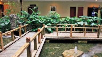 A wooden bridge over a pond with a fountain, surrounded by lush greenery and a wooden deck.